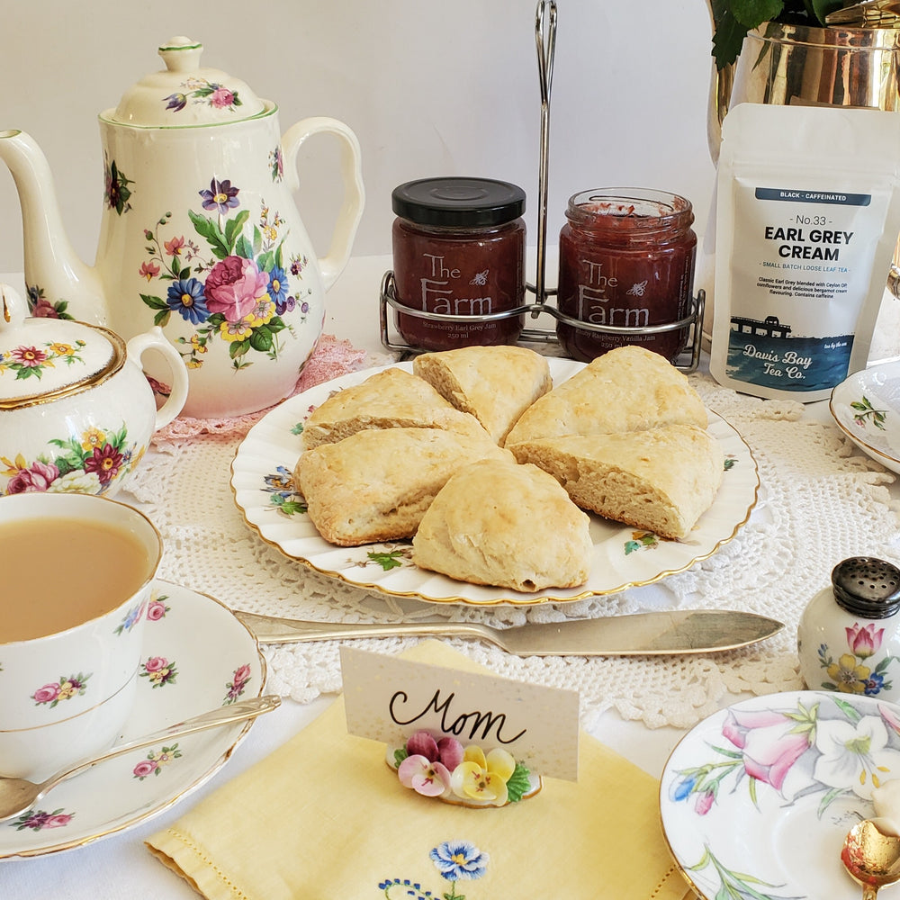 teapot and china tea cup filled with milky tea along with baked scones jam and tea package on a lovely decorated table setting