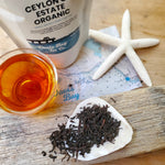 long wiry black tea leaves on seashell with cup of steeped tea, driftwood, starfish, map and bag of tea in background