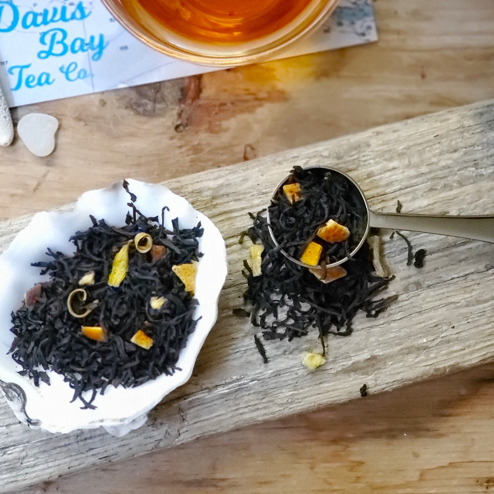 black tea with orange peel, orange blossoms on a seashell with a tea scoop, driftwood and part of tea cup with brewed tea in background